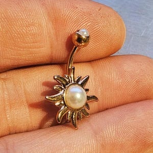 Könnte beinhalten: Ein goldfarbener Bauchnabelpiercing mit Sonnenmuster. Der Ring hat einen gebogenen Steg mit einer kleinen Perle oben und einer größeren Perle in der Mitte des Sonnenmotivs. Die Sonne hat spitze Strahlen.