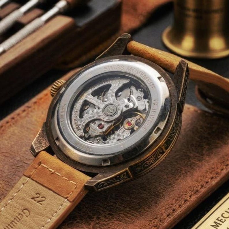 May include: A close-up of a vintage wristwatch with a visible mechanical movement. The watch has a bronze-coloured case, a light brown leather strap, and the text "SWISS MOVEMENT". The watch is displayed on a brown leather surface.