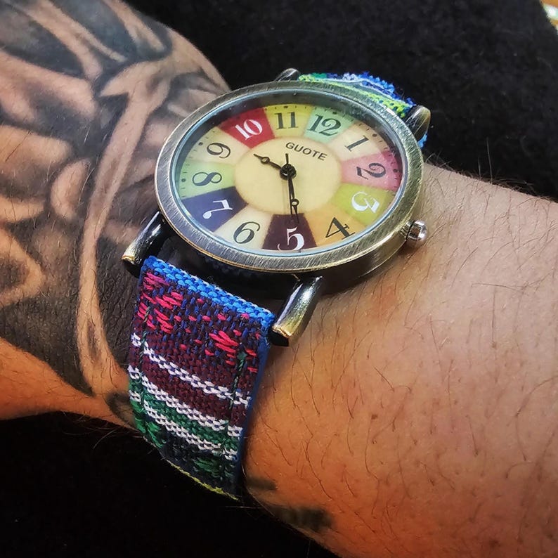 May include: A colorful analog wristwatch with a multi-colored face and a woven fabric band. The watch face has a rainbow design with numbers 1 through 12 and the word "QUOTE" printed on it.