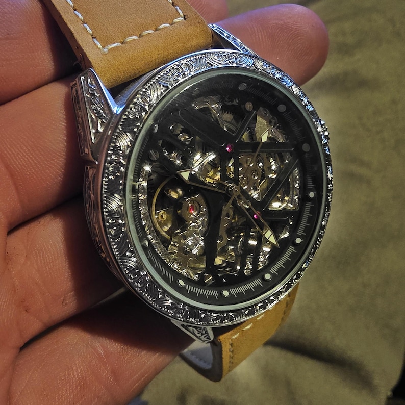 May include: A silver skeleton watch with a tan leather strap. The black watch face displays the internal mechanisms. The bezel and case feature detailed engravings. The strap is approximately 2.2 cm wide.