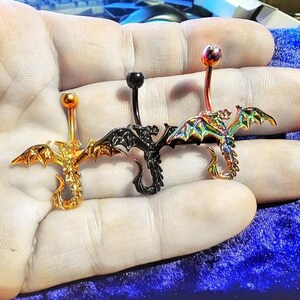 May include: Three dragon-shaped belly button rings in gold, black, and iridescent finishes. Each ring features a curved barbell with a decorative dragon pendant. The dragons have detailed wings and tails, adding a fantasy element to the jewelry.