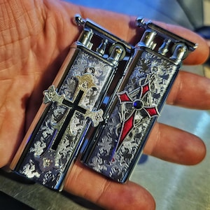 Könnte beinhalten: Zwei silberne Feuerzeuge mit verzierten Kreuzdesigns. Ein Kreuz ist silberfarben, das andere rot mit einem blauen Edelstein. Die Feuerzeuge haben ein detailliertes, graviertes Muster und ein metallisches Finish.