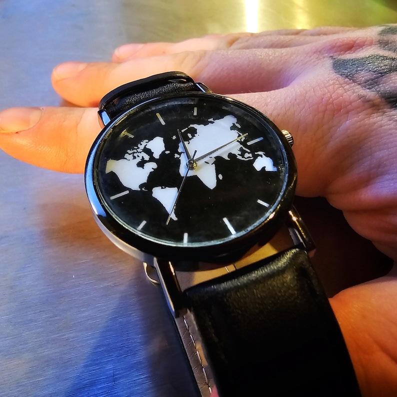 Puede incluir: Un reloj de pulsera negro con una correa de cuero negro. La esfera del reloj tiene un dise&ntilde;o de mapa del mundo blanco.