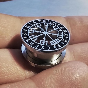 May include: A silver-colored ear gauge with a black and white design. The design features a circular pattern with runes and symbols. The gauge is held in a hand.