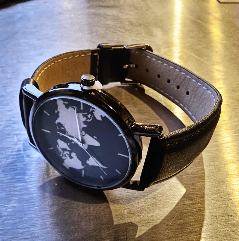 Puede incluir: Un reloj de pulsera de cuero negro con una esfera negra y un dise&ntilde;o de mapa del mundo blanco. El reloj tiene una hebilla plateada.