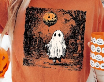 May include: Orange t-shirt featuring a Halloween-themed graphic of a ghost holding a pumpkin balloon in a graveyard setting. The shirt is paired with distressed jeans. A pumpkin mug and jack-o'-lantern are also visible.