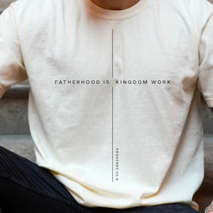 May include: Cream-colored t-shirt with the text "FATHERHOOD IS KINGDOM WORK" in black, centered on the chest. The shirt also features the Bible verse Proverbs 22:6. The shirt is paired with black pants.