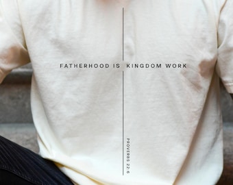 May include: Cream-colored t-shirt with the text "FATHERHOOD IS KINGDOM WORK" in black, centered on the chest. The shirt also features the Bible verse Proverbs 22:6. The shirt is paired with black pants.
