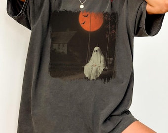 May include: Dark grey t-shirt featuring a spooky graphic of a ghost on a swing set under a full orange moon. The design includes a house and bats, creating a Halloween theme. The shirt has short sleeves and a relaxed fit.