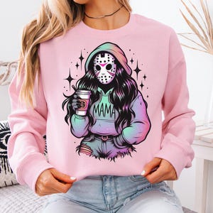 May include: Pink sweatshirt featuring a graphic of a masked figure with long black hair, holding a coffee cup. The word "MAMA" is printed on the figure's hoodie. The design has a colorful, gradient effect and is surrounded by star accents.