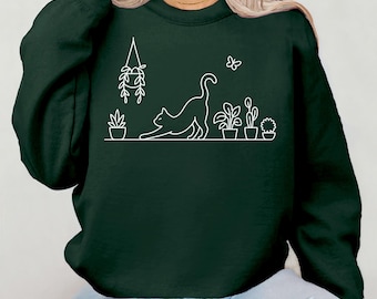 Peut inclure: Sweat-shirt vert foncé avec un dessin au trait blanc d'un chat s'étirant devant des plantes en pot. Une plante suspendue et un papillon sont également représentés. Le sweat-shirt est de couleur unie et à manches longues.
