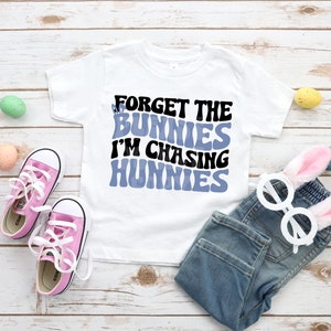 May include: White t-shirt with blue and black text that reads "Forget the bunnies I'm chasing hunnies". The shirt is paired with blue jeans, pink sneakers, and bunny ears.