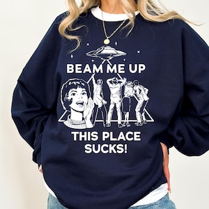 May include: Navy blue sweatshirt with a white graphic that reads "BEAM ME UP THIS PLACE SUCKS!" The graphic features a retro illustration of a woman, a UFO, and figures being beamed up. The sweatshirt has a white crew neck.