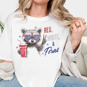 May include: White t-shirt featuring a raccoon wearing red, white, and blue sunglasses, holding a red cup with a straw. The text "Red, White, & Feral" is printed on the shirt in red and blue.
