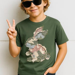 May include: Olive green t-shirt featuring a camouflage bunny design. The bunny is made of patches of fabric in shades of green, brown, and blue. The shirt has short sleeves and a crew neck.