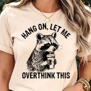 May include: Beige t-shirt featuring a black and white illustration of a raccoon holding a coffee cup. The text "HANG ON, LET ME OVERTHINK THIS" is printed above and below the raccoon graphic. The shirt has short sleeves.