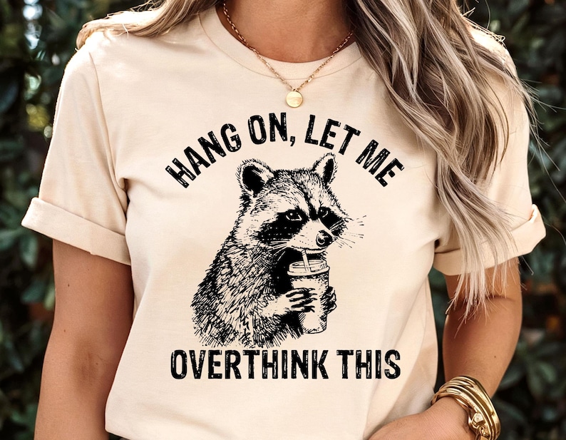 May include: Beige t-shirt featuring a black and white illustration of a raccoon holding a coffee cup. The text "HANG ON, LET ME OVERTHINK THIS" is printed above and below the raccoon graphic. The shirt has short sleeves.