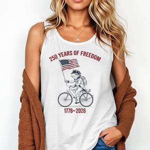 May include: White tank top featuring a patriotic design. The graphic depicts a frog riding a bicycle and holding an American flag. Text above reads "250 YEARS OF FREEDOM" with the years "1776-2026" below the bicycle.
