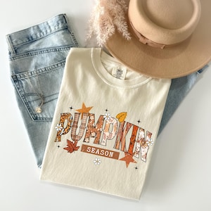 Pu&ograve; includere: T-shirt color crema con la scritta "PUMPKIN SEASON" in un design a tema autunnale, con motivi arancioni, marroni e floreali. La maglietta &egrave; abbinata a jeans blu chiaro, un cappello color cachi e una delicata collana.