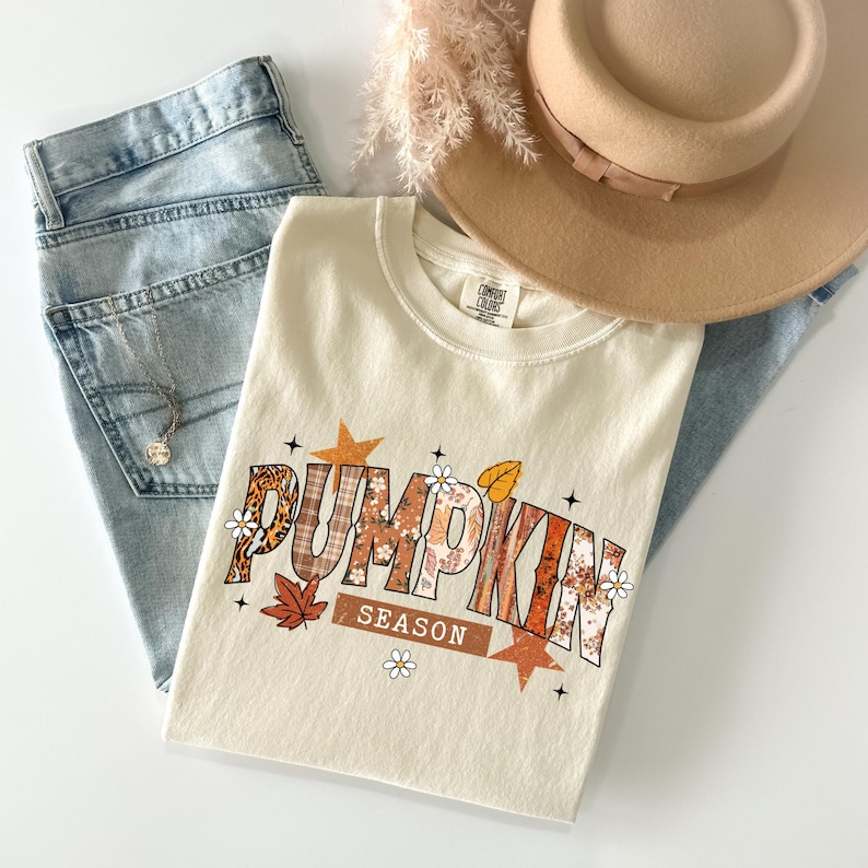Pu&ograve; includere: T-shirt color crema con la scritta "PUMPKIN SEASON" in un design a tema autunnale, con motivi arancioni, marroni e floreali. La maglietta &egrave; abbinata a jeans blu chiaro, un cappello color cachi e una delicata collana.