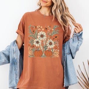 May include: Rust-colored t-shirt featuring a floral design with white daisies, orange flowers, and two pumpkins. The design is set against a square background. The shirt has rolled-up sleeves and a relaxed fit.