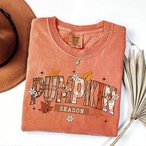 Pu&ograve; includere: Una maglietta Comfort Colors color corallo con le parole "PUMPKIN SEASON" in un design a tema autunnale. Le lettere sono riempite con stampa leopardata, scozzese e motivi floreali. La maglietta &egrave; accessoria con una collana d'oro e un cappello marrone.