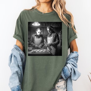 Puede incluir: Camiseta verde con una imagen en blanco y negro de dos figuras con delantales, una de ellas con una m&aacute;scara de hockey, horneando en una cocina. La imagen est&aacute; centrada en la camiseta, ofreciendo un dise&ntilde;o &uacute;nico y tem&aacute;tico. La camiseta es c&oacute;moda e informal.