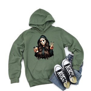 May include: An olive green hooded sweatshirt featuring a graphic of a person in a hockey mask, holding a coffee cup, and making a peace sign. Black and white Converse trainers and blue jeans are also in the image.
