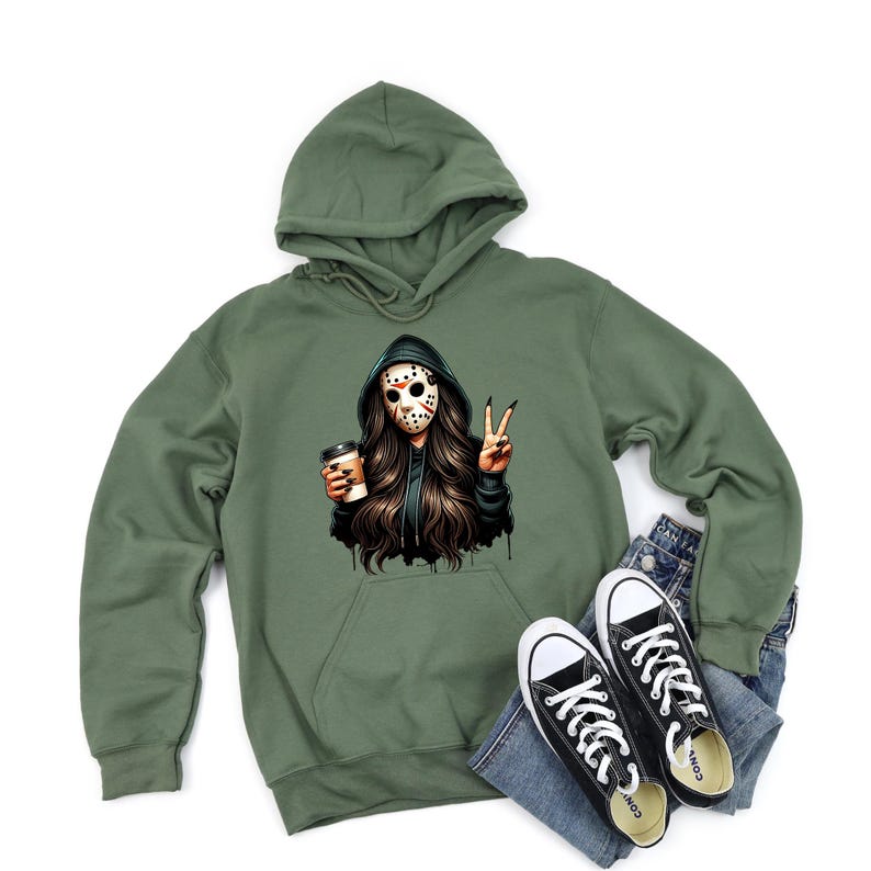 May include: An olive green hooded sweatshirt featuring a graphic of a person in a hockey mask, holding a coffee cup, and making a peace sign. Black and white Converse trainers and blue jeans are also in the image.