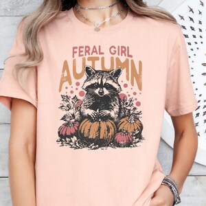 May include: A pale pink t-shirt with a raccoon illustration and pumpkins. The text "FERAL GIRL AUTUMN" is printed above the raccoon. The shirt has short sleeves and a round neck.