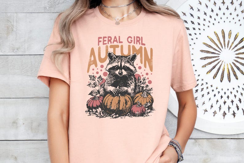 May include: A pale pink t-shirt with a raccoon illustration and pumpkins. The text "FERAL GIRL AUTUMN" is printed above the raccoon. The shirt has short sleeves and a round neck.