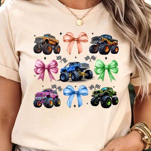 Puede incluir: Camiseta beige con un dise&ntilde;o de seis monster trucks coloridos y seis lazos en tonos rosa, naranja, azul y verde. Cada cami&oacute;n tiene neum&aacute;ticos grandes y est&aacute; decorado con banderas de carreras.