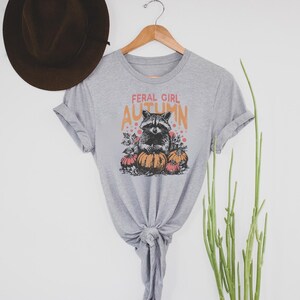May include: A grey t-shirt featuring the words "FERAL GIRL AUTUMN" and a raccoon illustration with pumpkins. The t-shirt is displayed on a wooden hanger.