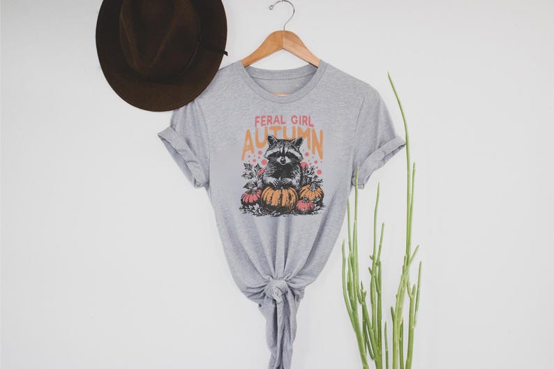 May include: A grey t-shirt featuring the words "FERAL GIRL AUTUMN" and a raccoon illustration with pumpkins. The t-shirt is displayed on a wooden hanger.