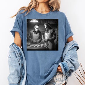 Puede incluir: Una camiseta azul con una imagen en blanco y negro de dos figuras con delantales, una de ellas con una m&aacute;scara de hockey. La imagen representa una escena de cocina con las figuras preparando comida. La camiseta tiene mangas cortas y un corte holgado.