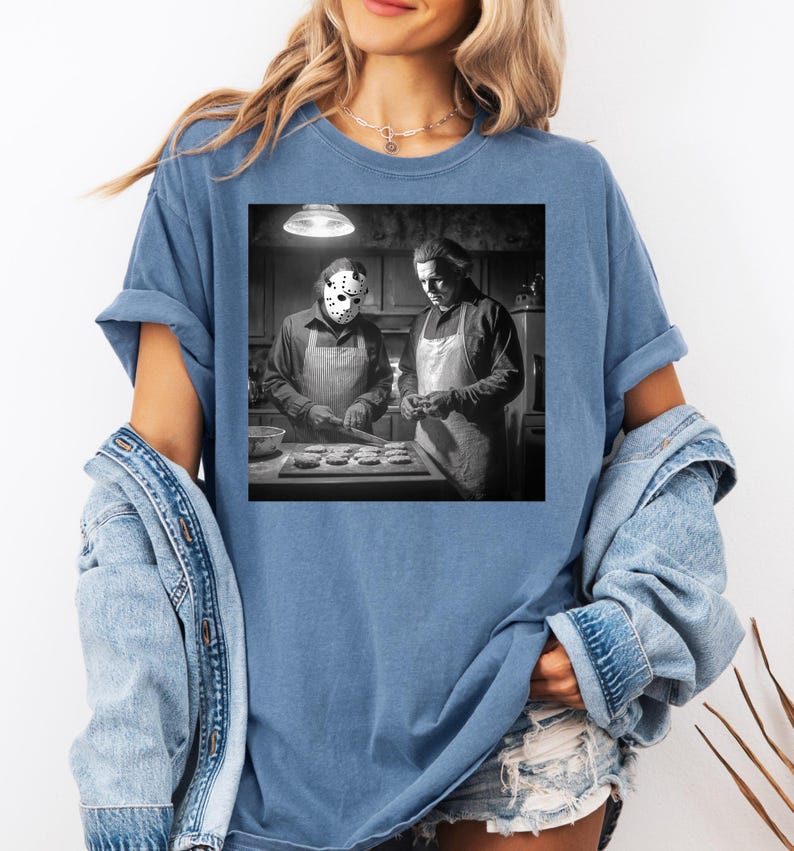 Puede incluir: Una camiseta azul con una imagen en blanco y negro de dos figuras con delantales, una de ellas con una m&aacute;scara de hockey. La imagen representa una escena de cocina con las figuras preparando comida. La camiseta tiene mangas cortas y un corte holgado.