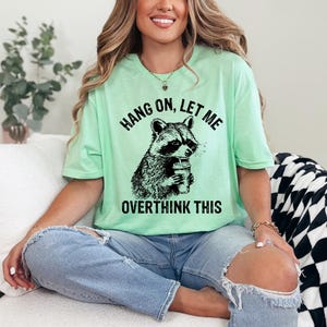 May include: A light green t-shirt with the text "HANG ON, LET ME OVERTHINK THIS" and a black and white graphic of a raccoon holding a cup. The shirt is worn with blue jeans.
