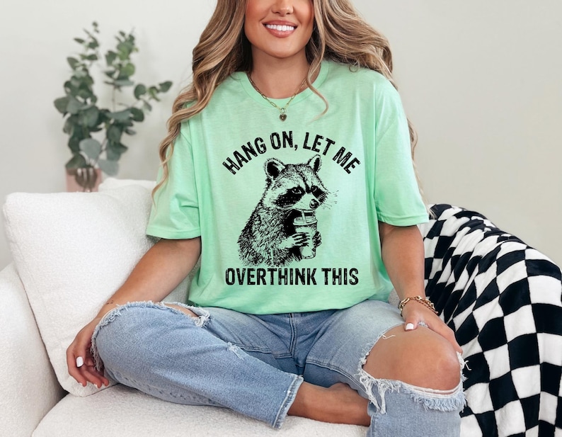 May include: A light green t-shirt with the text "HANG ON, LET ME OVERTHINK THIS" and a black and white graphic of a raccoon holding a cup. The shirt is worn with blue jeans.
