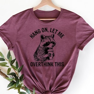 May include: Burgundy t-shirt with a raccoon graphic holding a drink. The text above the raccoon reads "Hang On, Let Me" and below it says "Overthink This". The shirt is hanging on a wooden hanger.