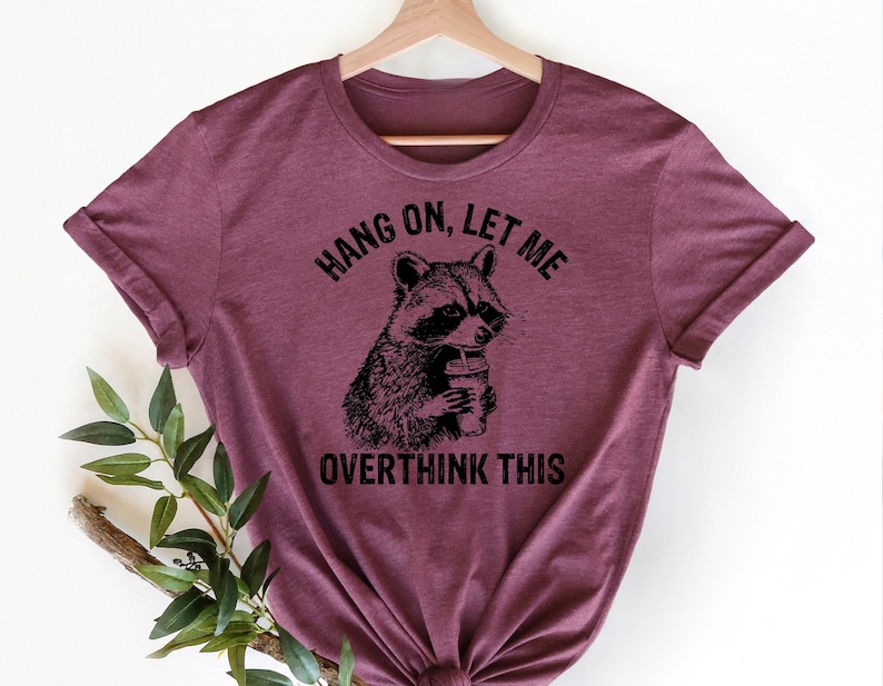 May include: Burgundy t-shirt with a raccoon graphic holding a drink. The text above the raccoon reads "Hang On, Let Me" and below it says "Overthink This". The shirt is hanging on a wooden hanger.