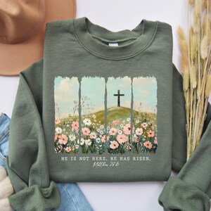 May include: An olive green sweatshirt featuring a floral and cross design. The graphic includes a cross on a hill, surrounded by flowers, with the text "HE IS NOT HERE. HE HAS RISEN. Matthew 28:6".