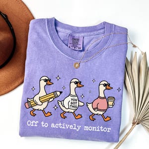 May include: A light purple t-shirt with the phrase "Off to actively monitor" and a graphic of three cartoon ducks wearing sunglasses. One duck holds a pencil, another a shirt that says "You Got This", and the third a coffee cup.