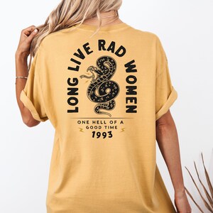 Peut inclure: T-shirt jaune moutarde avec un motif noir de serpent et le texte "LONG LIVE RAD WOMEN" et "ONE HELL OF A GOOD TIME 1993". Le t-shirt a des manches courtes et une coupe d&eacute;contract&eacute;e.