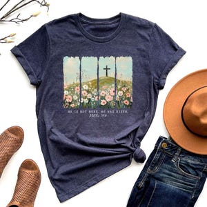 May include: Navy blue t-shirt featuring a graphic of a cross on a hill with flowers, and the text "HE IS NOT HERE. HE HAS RISEN. Matthew 28:6". The shirt is styled with a brown hat, jeans, and brown sandals.