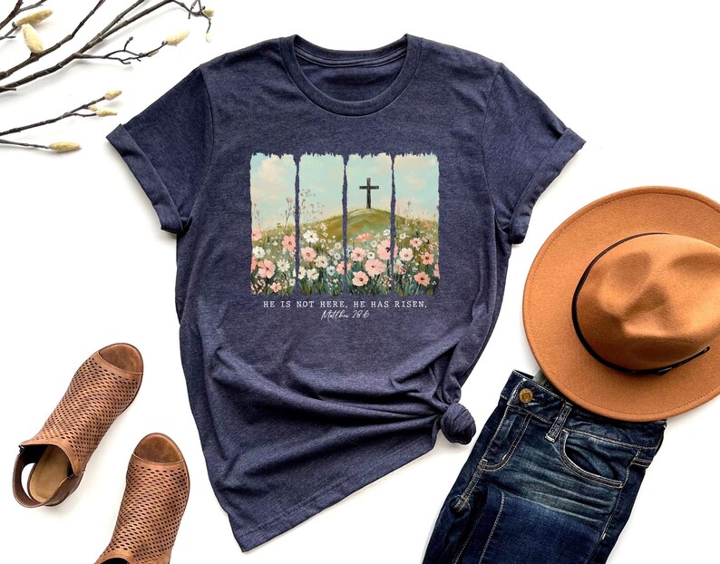 May include: Navy blue t-shirt featuring a graphic of a cross on a hill with flowers, and the text "HE IS NOT HERE. HE HAS RISEN. Matthew 28:6". The shirt is styled with a brown hat, jeans, and brown sandals.