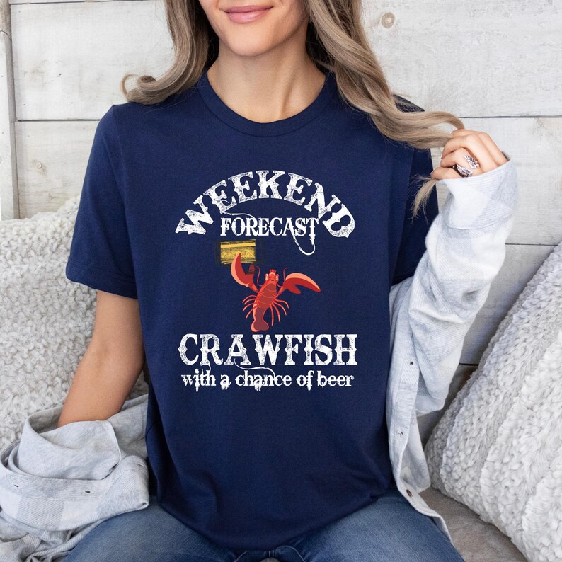 May include: A navy blue t-shirt with a white graphic design that says "Weekend Forecast: Crawfish with a chance of beer". The graphic includes a cartoon crawfish holding a can of beer.