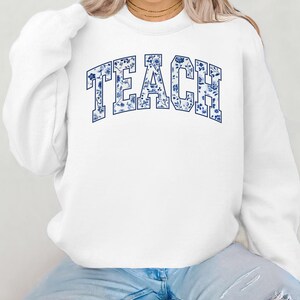 May include: White sweatshirt with a blue floral pattern spelling out the word "TEACH" in a varsity style font.