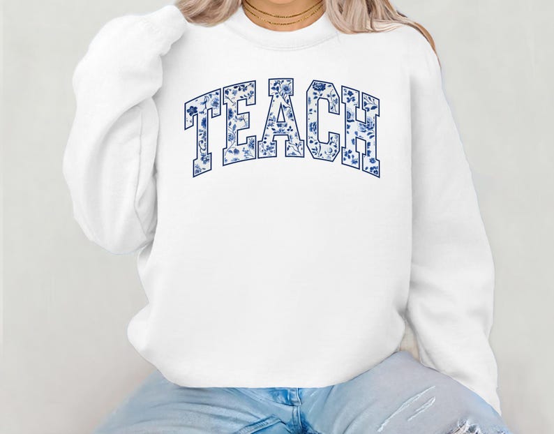 May include: White sweatshirt with a blue floral pattern spelling out the word "TEACH" in a varsity style font.