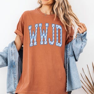 May include: A rust-colored t-shirt with the letters "WWJD" in a blue floral pattern. The shirt has short sleeves and a relaxed fit. A light blue denim jacket is worn over the shirt.