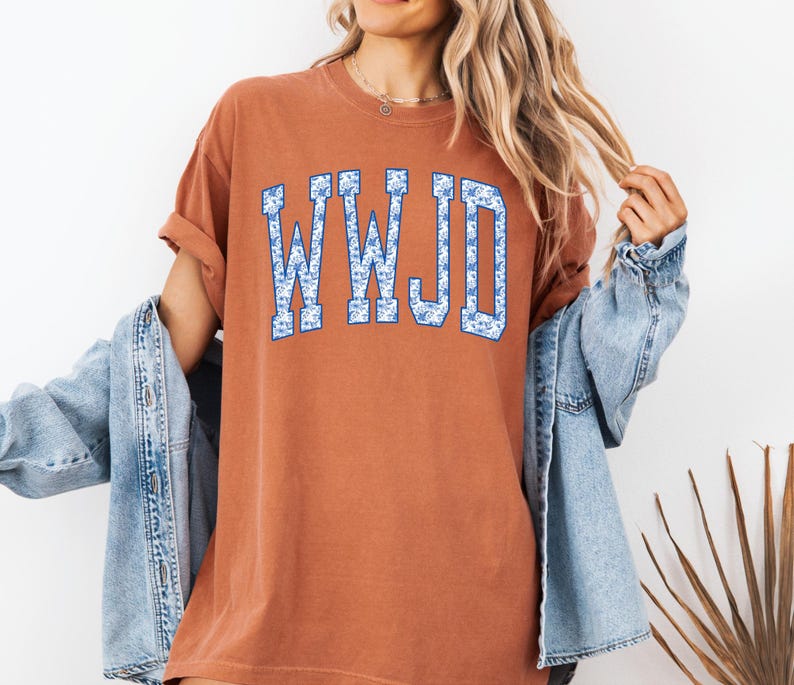 May include: A rust-colored t-shirt with the letters "WWJD" in a blue floral pattern. The shirt has short sleeves and a relaxed fit. A light blue denim jacket is worn over the shirt.